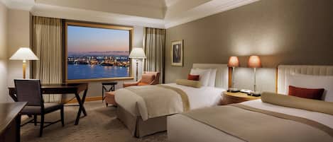 A hotel room with a large window offering a city view, a desk with a chair, two beds, and two bedside tables with lamps.