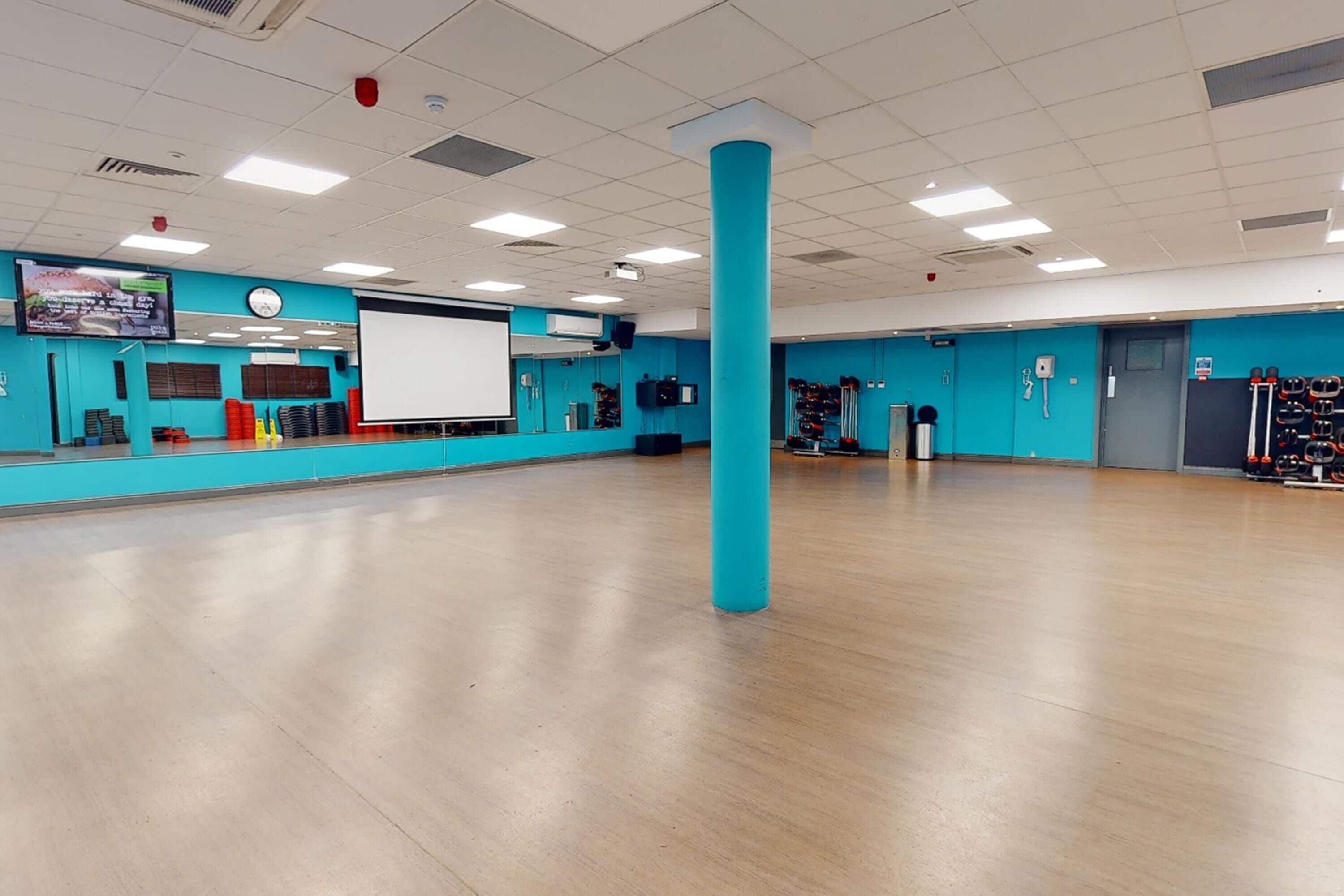 A spacious dance studio with a wooden floor, a large blue pillar, a projection screen, and exercise equipment.