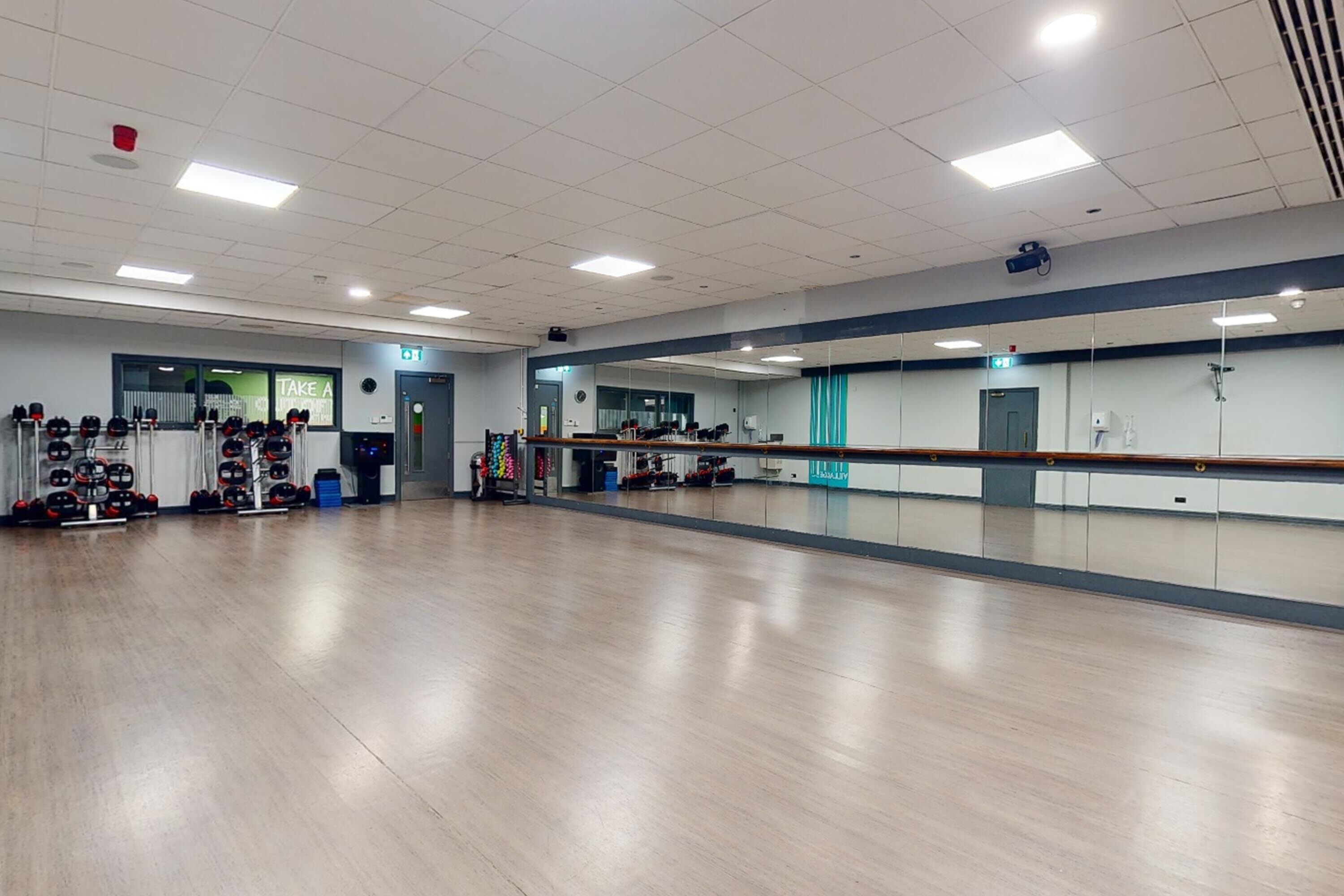 A spacious room with a long mirror, wooden flooring, and a row of lockers on the left.