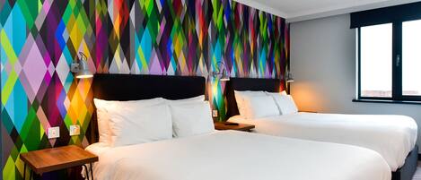 A hotel room with two beds, a colorful diamond-patterned wall, and a window with a view.