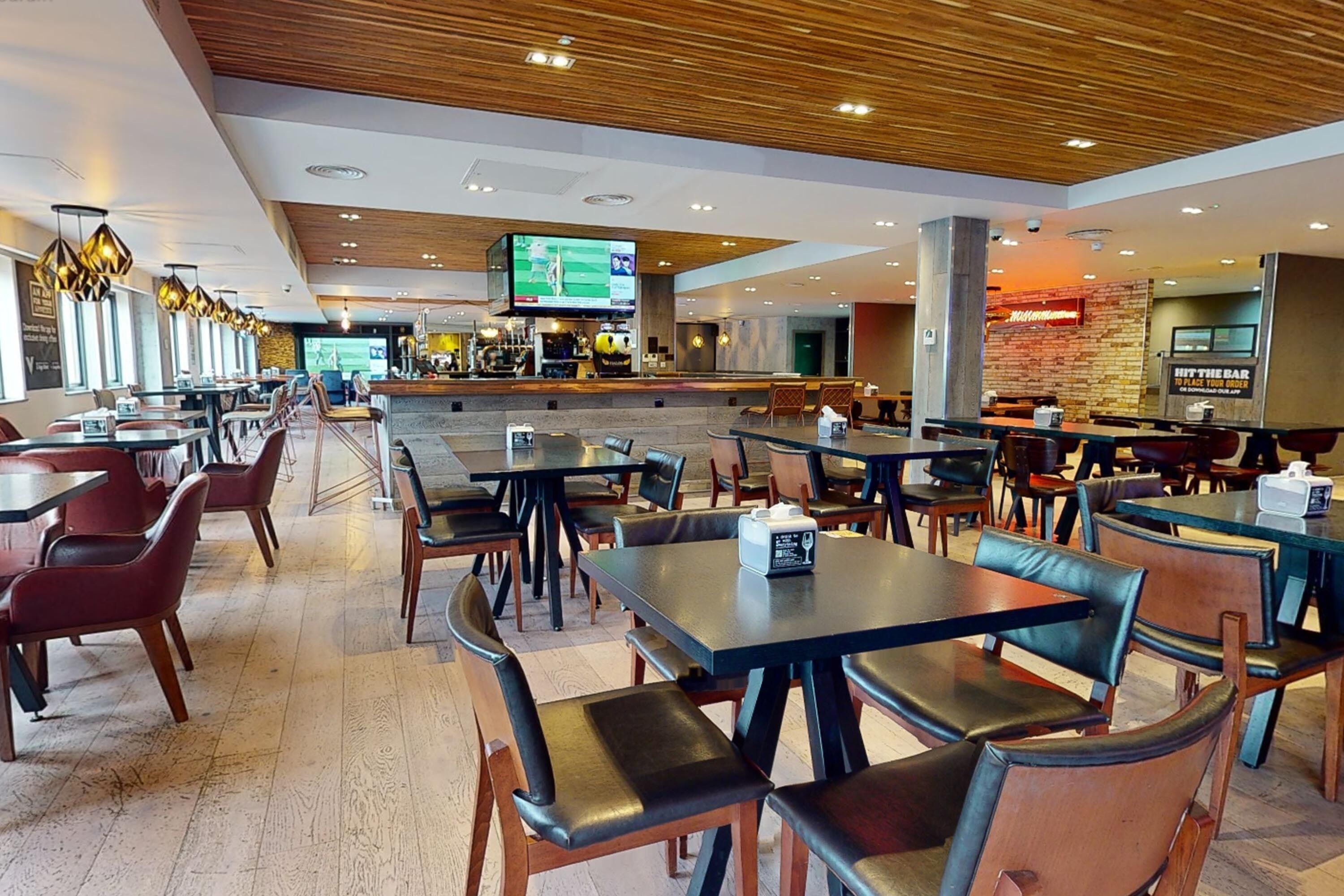 A modern restaurant with multiple dining tables and booths, a bar area, and a television screen displaying sports.