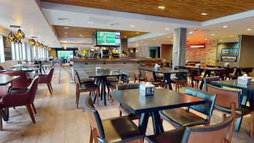 A modern restaurant with multiple dining tables and booths, a bar area, and a television screen displaying sports.