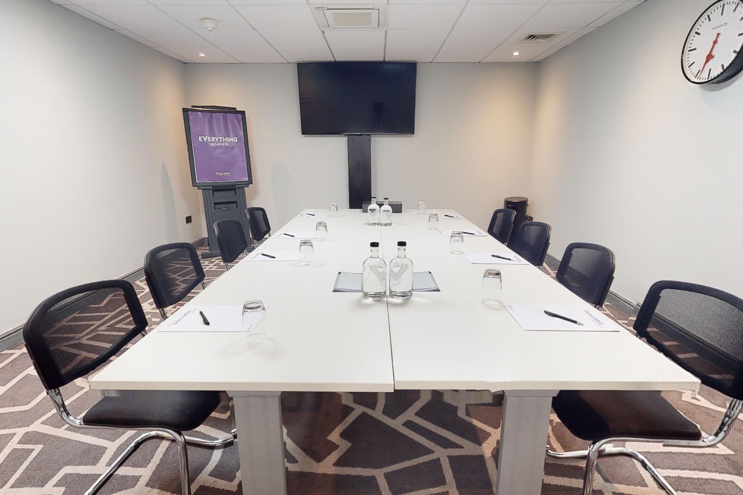 A conference room with a long table, chairs, a TV screen, and a clock on the wall.