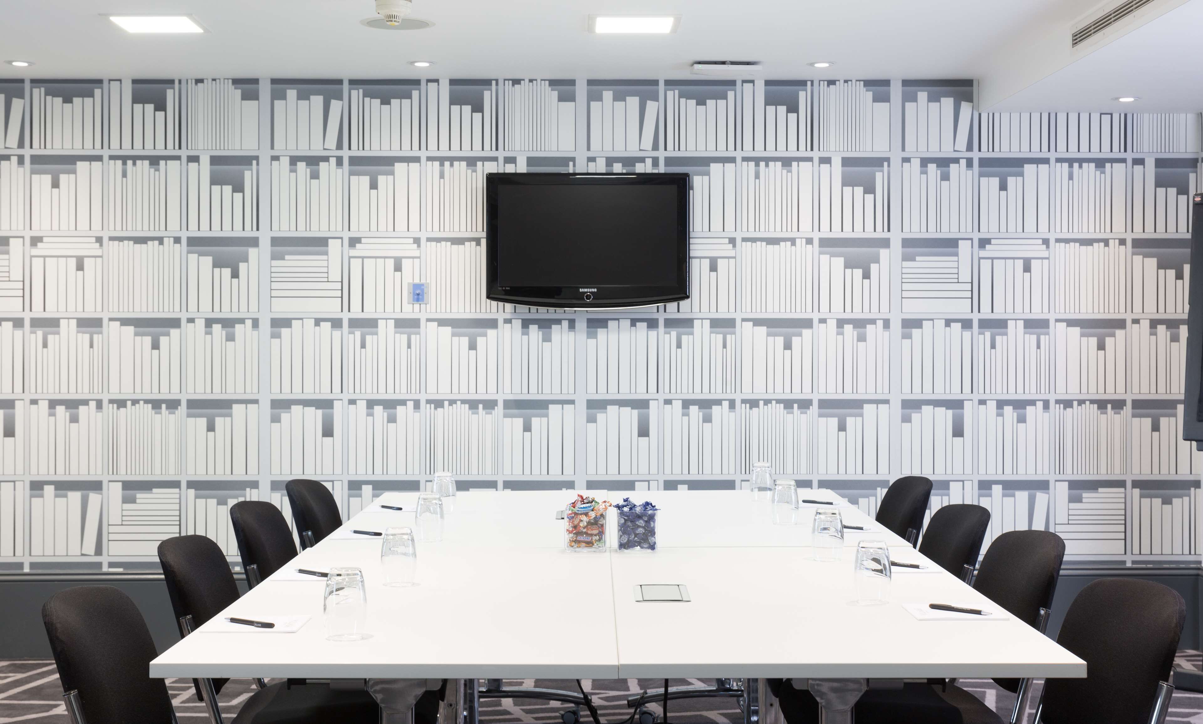 A conference room with a white table, black chairs, a Samsung TV mounted on the wall, and a bookshelf in the background.