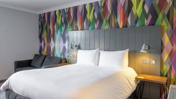 Quarto de hotel moderno com uma cama grande, um sofá cinza estofado, uma parede com padrão geométrico colorido e uma pequena mesa lateral de madeira.