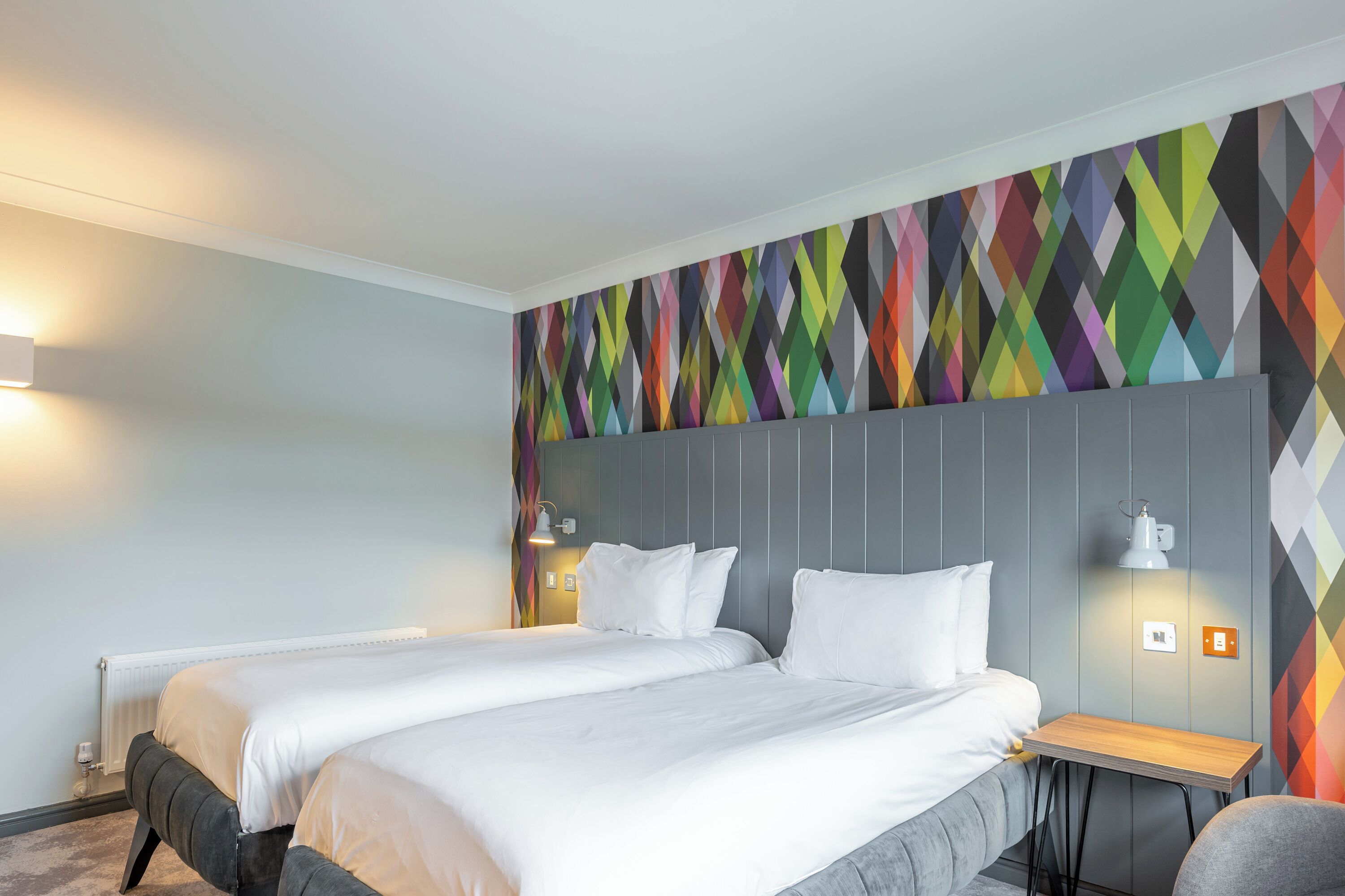 Ein Hotelzimmer mit zwei Betten, einem kleinen Holztisch und einem farbenfrohen geometrischen Wanddesign.