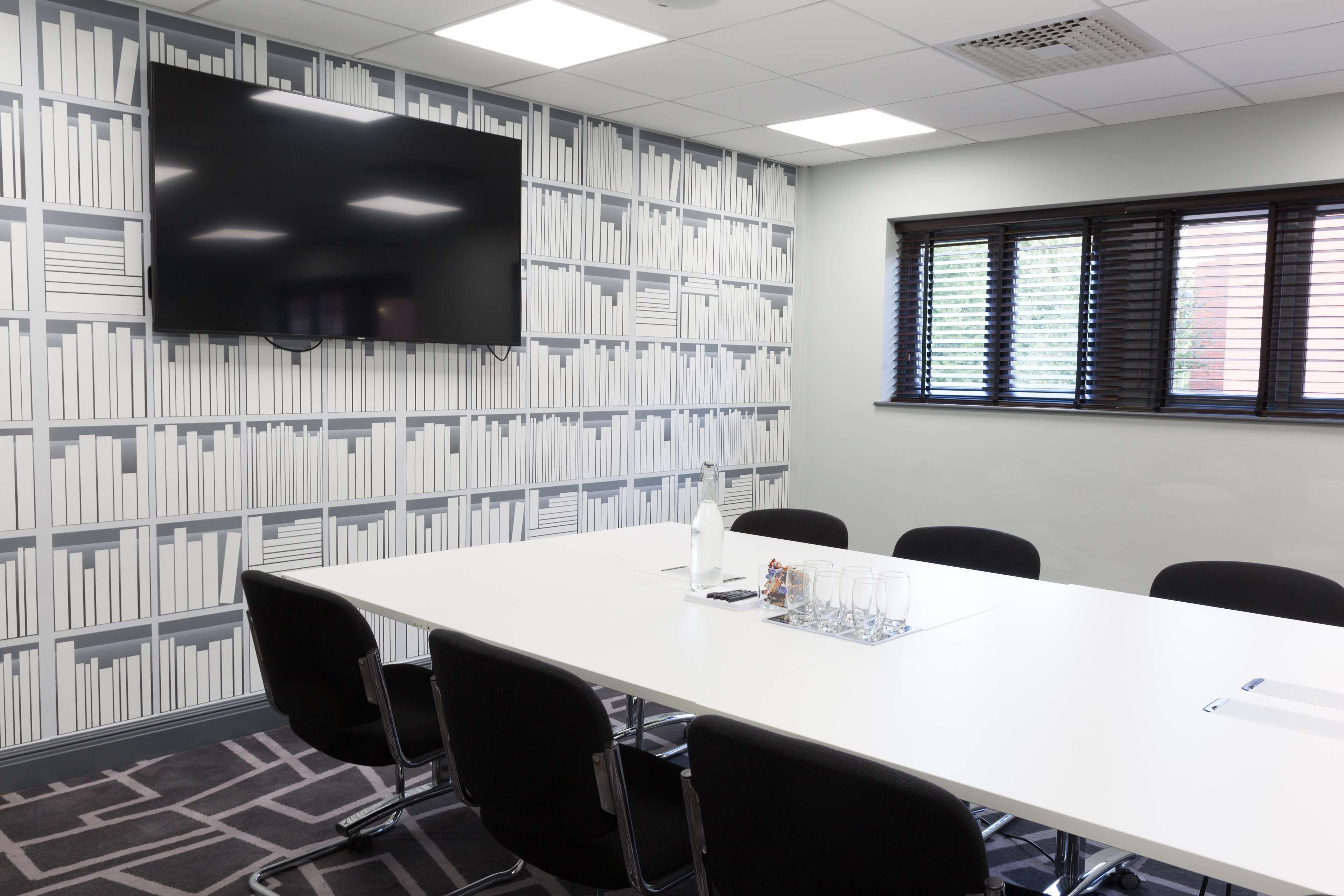 A conference room with a long white table, black chairs, a flat-screen TV mounted on the wall, and a bookshelf with books.