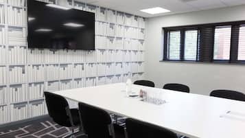 A conference room with a long white table, black chairs, a flat-screen TV mounted on the wall, and a bookshelf with books.