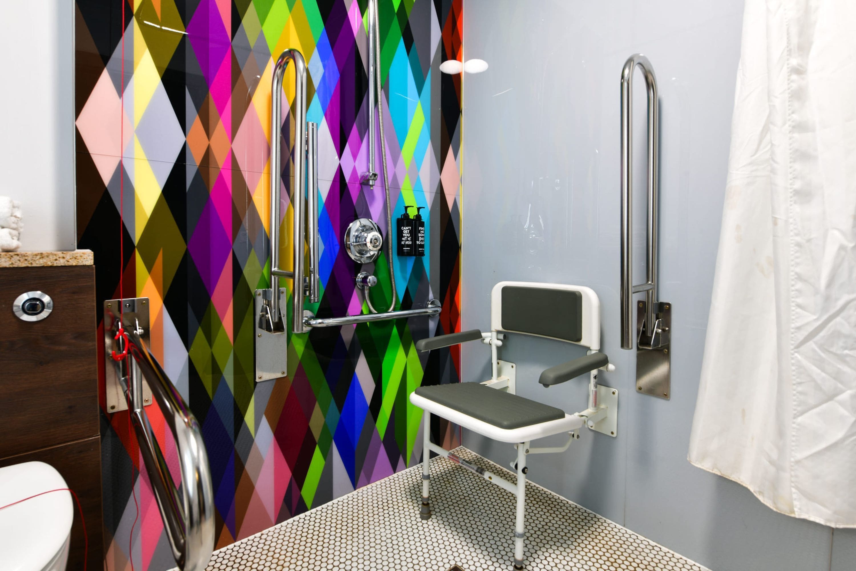 Casa de banho com parede em padrão de losangos coloridos, chuveiro com chuveiro manual, sanita e barra de apoio.