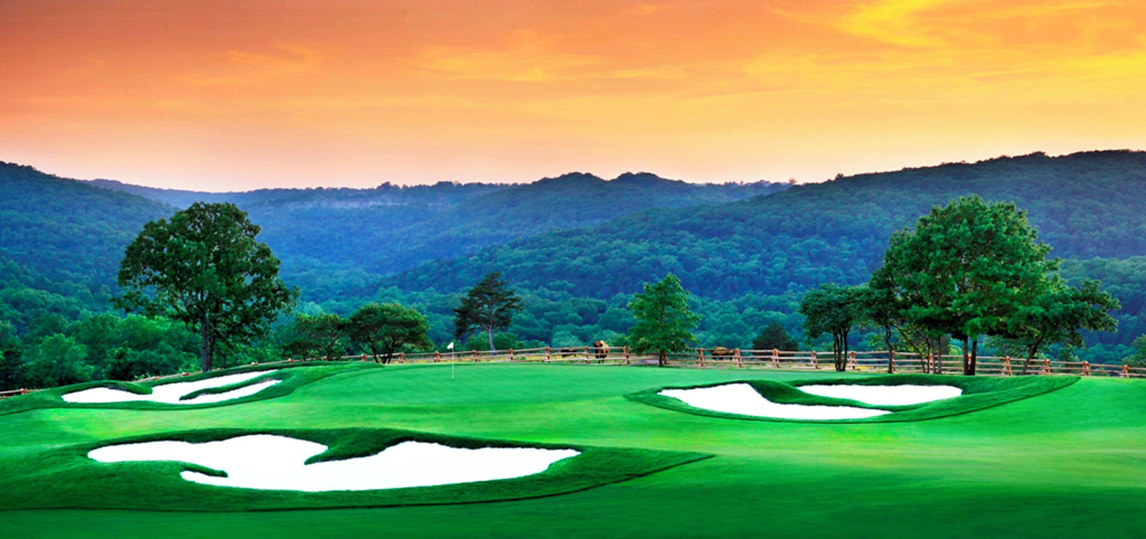 Campo de golf con búnkeres de arena, árboles y montañas al fondo.