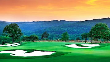 Campo de golf con búnkeres de arena, árboles y montañas al fondo.