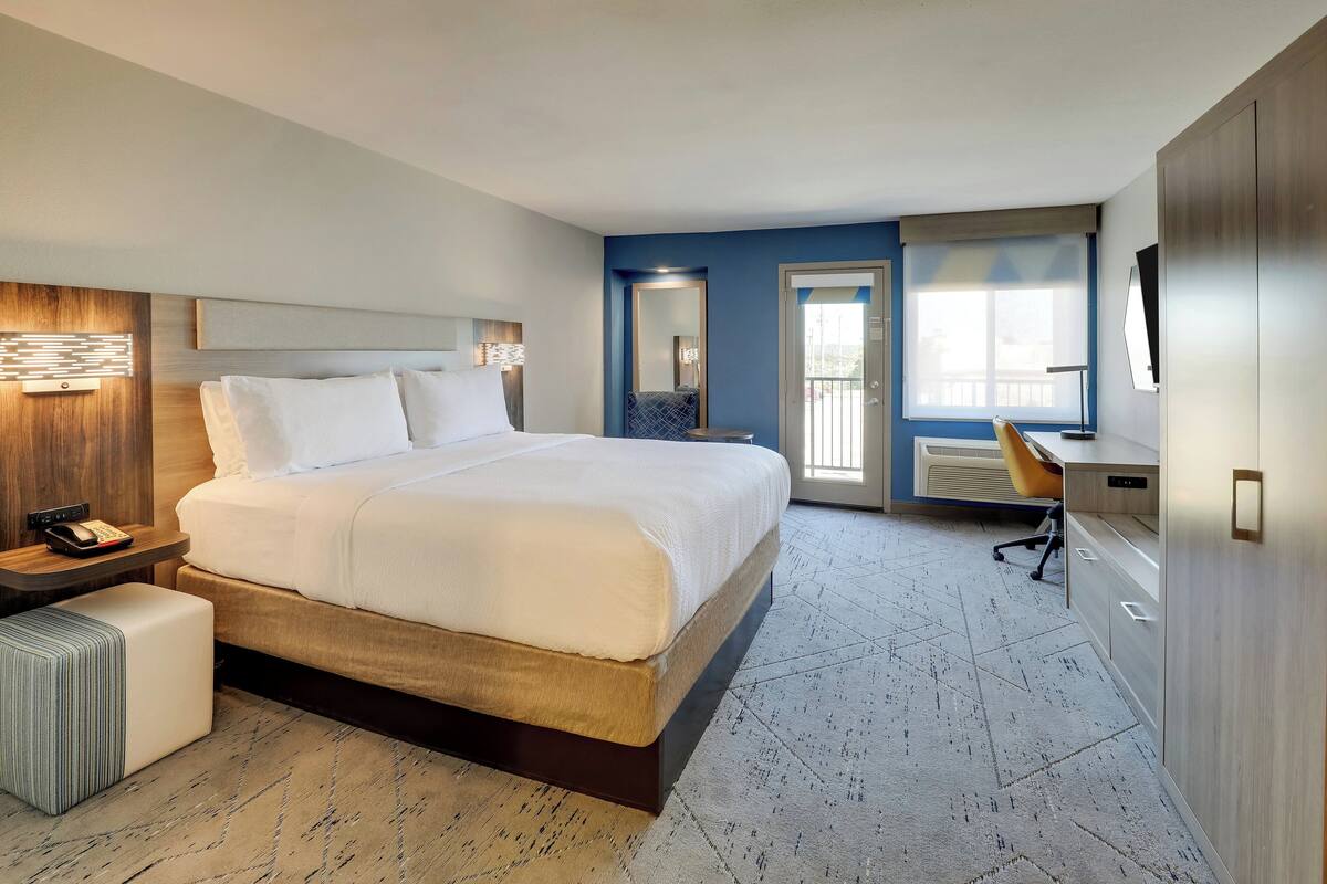 A modern hotel room with a large bed, bedside table with a phone, a desk with a chair, and a large window with a view.