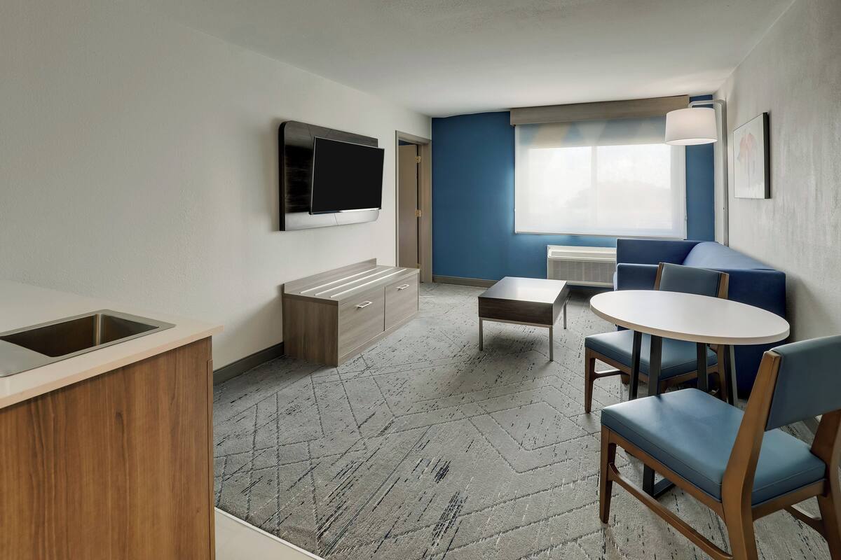 A modern hotel room with a kitchenette, a flat-screen TV, a sofa, and a dining table.