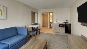 A modern living room with a blue sofa, a wooden coffee table, and a dining area with a small table and chairs. A kitchen with a microwave and sink is visible in the background. - Holiday Inn Express Hotel & Suites Branson 76 Central by IHG (Branson)