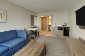 A modern living room with a blue sofa, a wooden coffee table, and a dining area with a small table and chairs. A kitchen with a microwave and sink is visible in the background. - Holiday Inn Express Hotel & Suites Branson 76 Central by IHG (Branson)