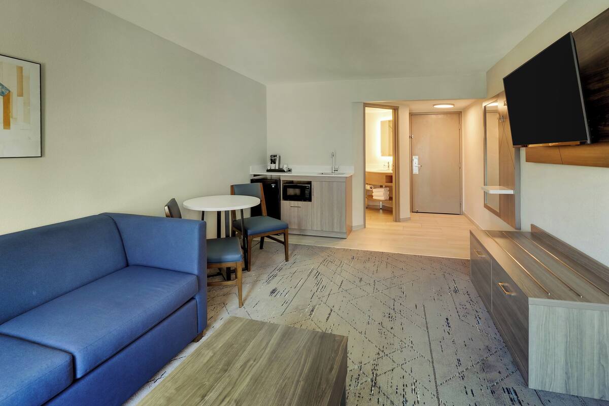 A compact living space with a sofa, a dining table, chairs, a kitchenette, and a TV mounted on the wall.