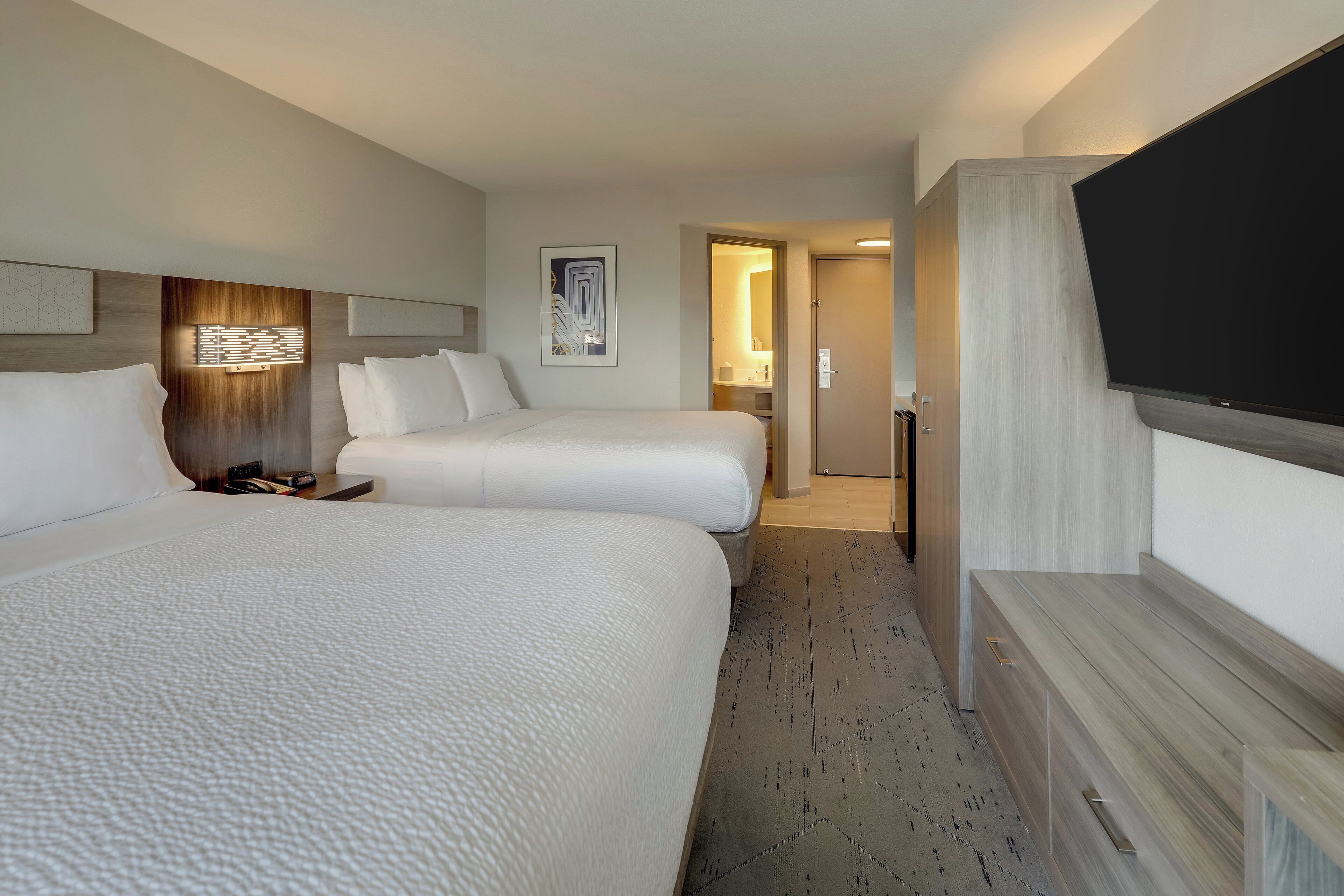 A hotel room with two beds, a flat-screen TV, a wooden headboard, a wall-mounted light, and a framed picture.