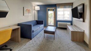 Ein kleines Zimmer mit einer Couch, einem Schreibtisch, einem kleinen Tisch, einem Fernseher und einem Fenster mit Jalousien.