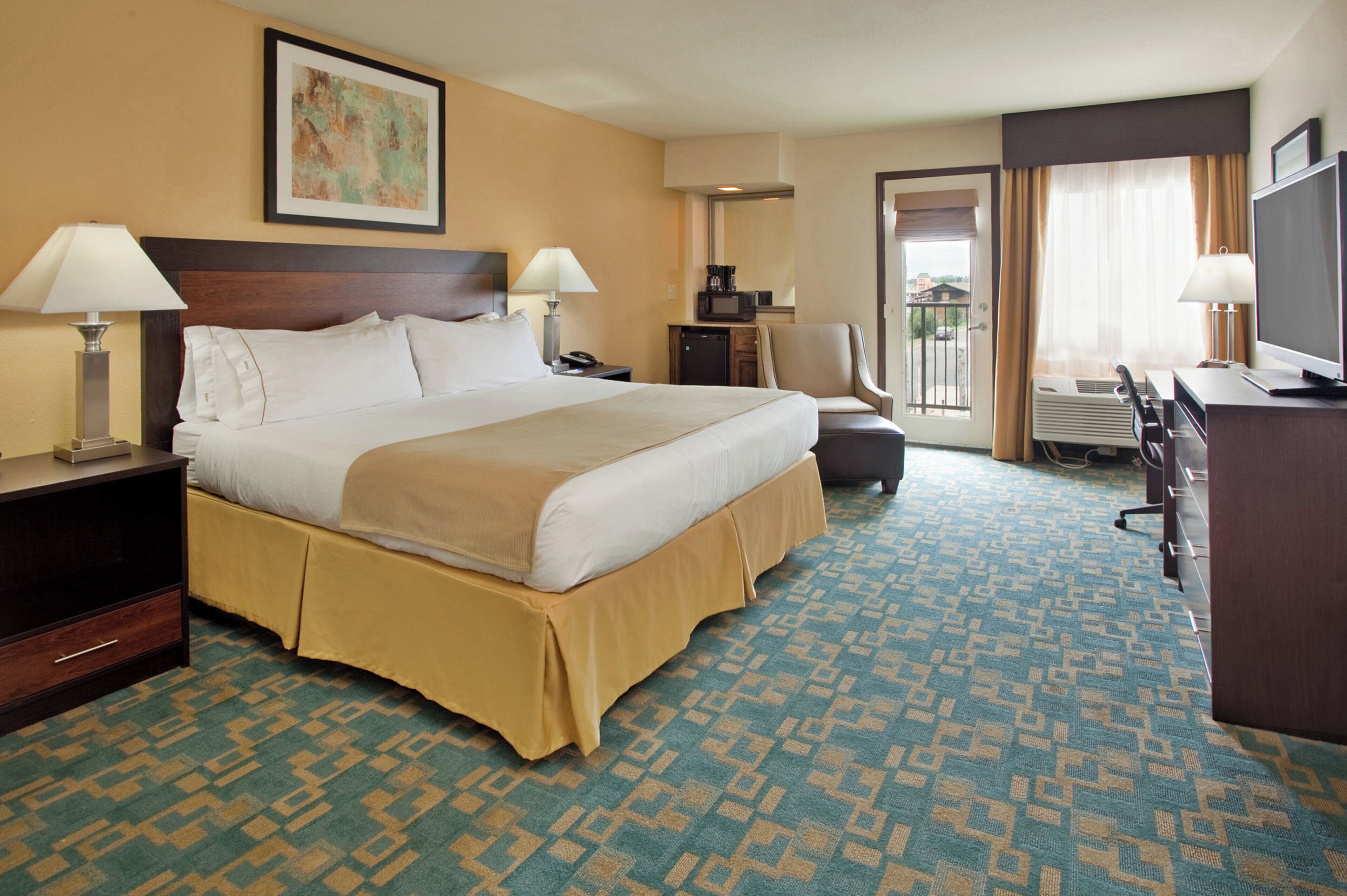 A hotel room with a large bed, two bedside lamps, a desk with a computer, a chair, a television, and a window with curtains.