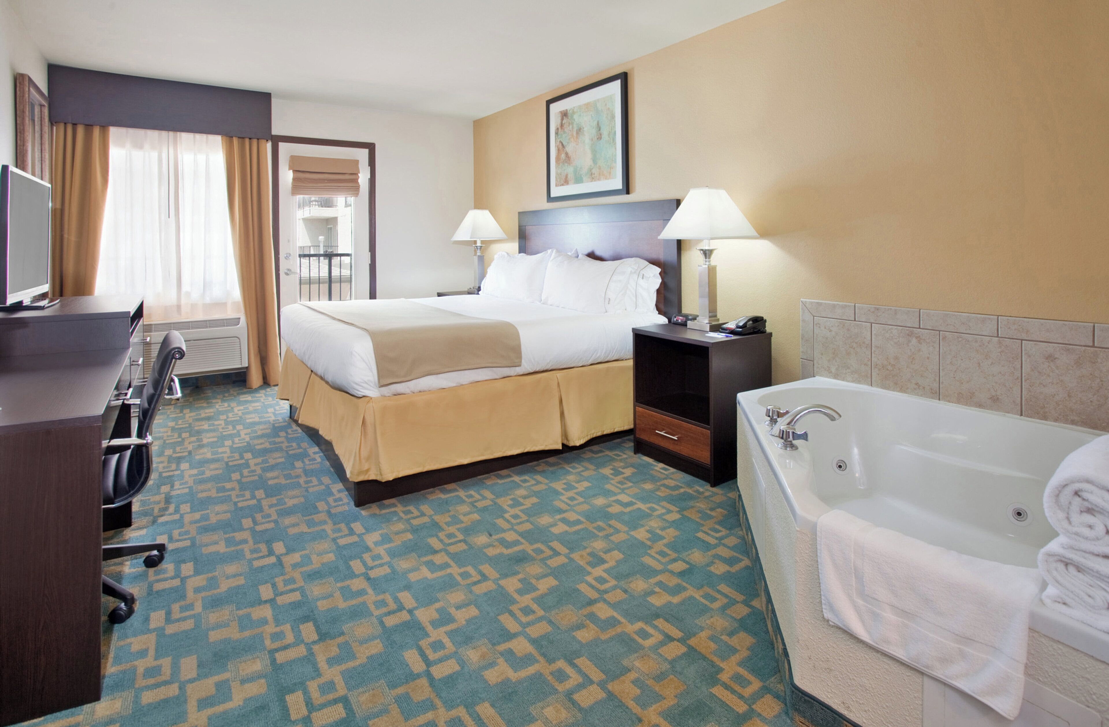 A hotel room with a bed, bedside tables, a desk, a chair, a bathroom with a bathtub, and a window with curtains.