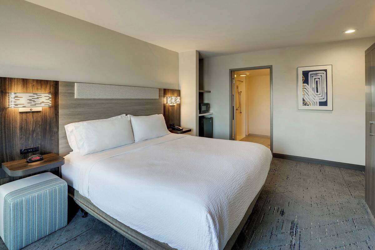 A modern hotel room with a large bed, bedside tables, a nightstand, a wall-mounted TV, and a bathroom door.