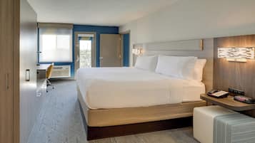 A modern hotel room with a large bed, bedside table with a phone and alarm clock, a small seating area, and a view of the outdoors.