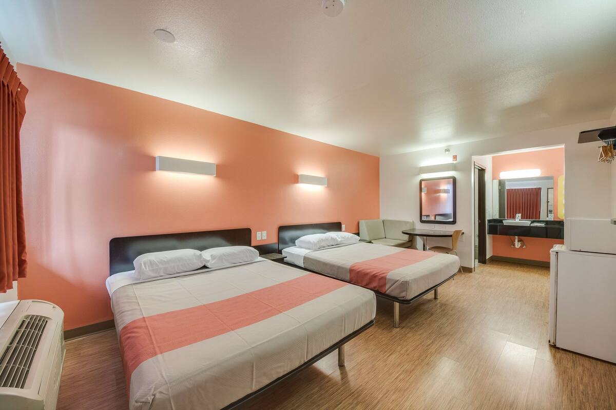 Une chambre d’hôtel avec deux lits, un bureau, un réfrigérateur et une salle de bain visible à travers une porte ouverte.