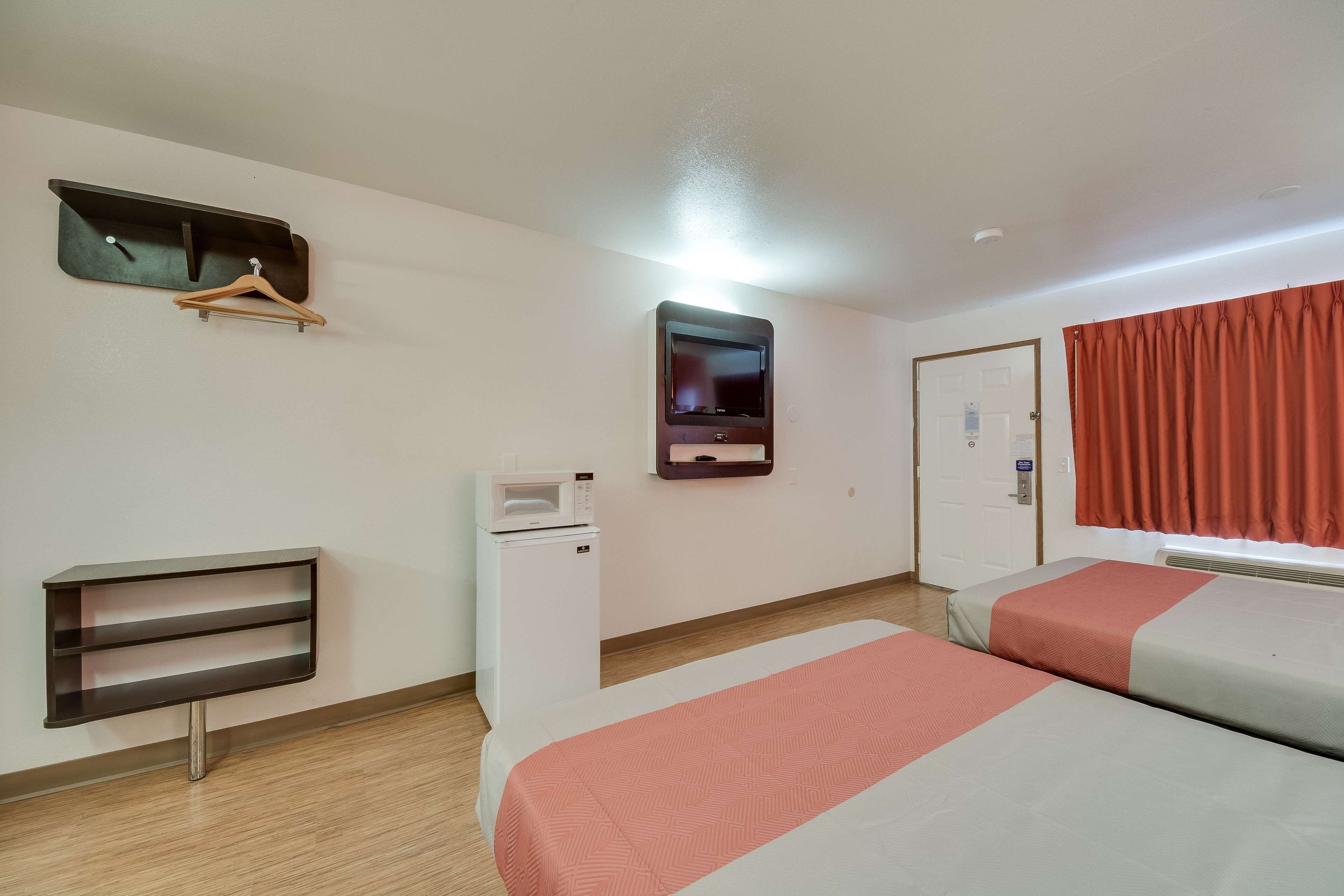 A hotel room with two beds, a TV mounted on the wall, a microwave, a small shelf, and a window with curtains.