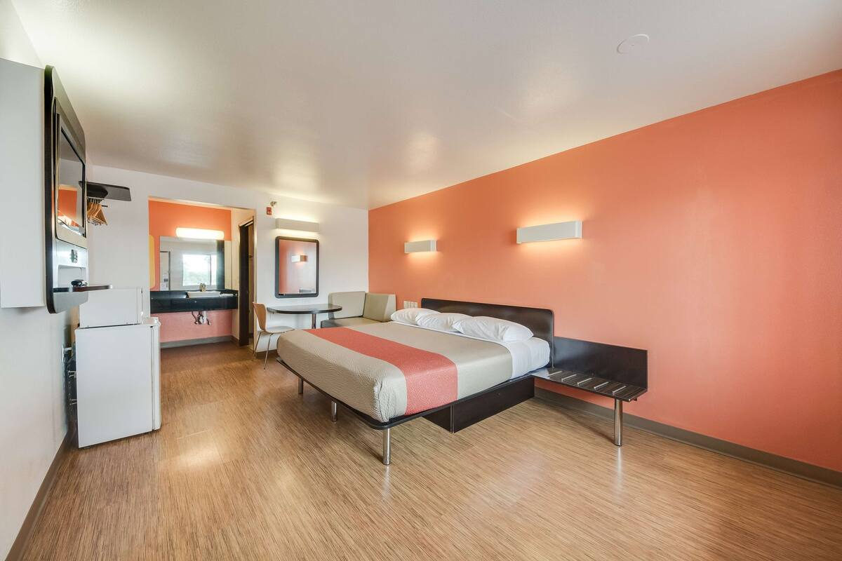 Une chambre d’hôtel équipée d’un lit, d’un bureau, d’une chaise, d’un réfrigérateur et d’une salle de bain, le tout visible à travers une porte ouverte.