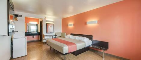 Habitación de hotel con cama, escritorio, silla, refrigerador y baño visible a través de una puerta abierta.