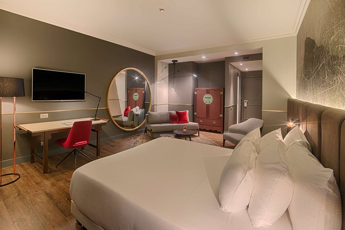 A hotel room with a large bed, a desk with a chair, a TV, a mirror, and a wardrobe.
