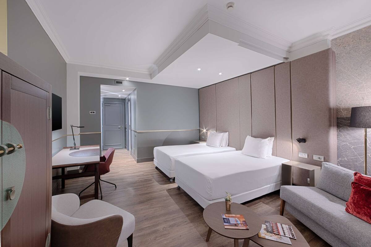 A hotel room with two beds, a desk, a chair, a sofa, and a small table with a magazine.