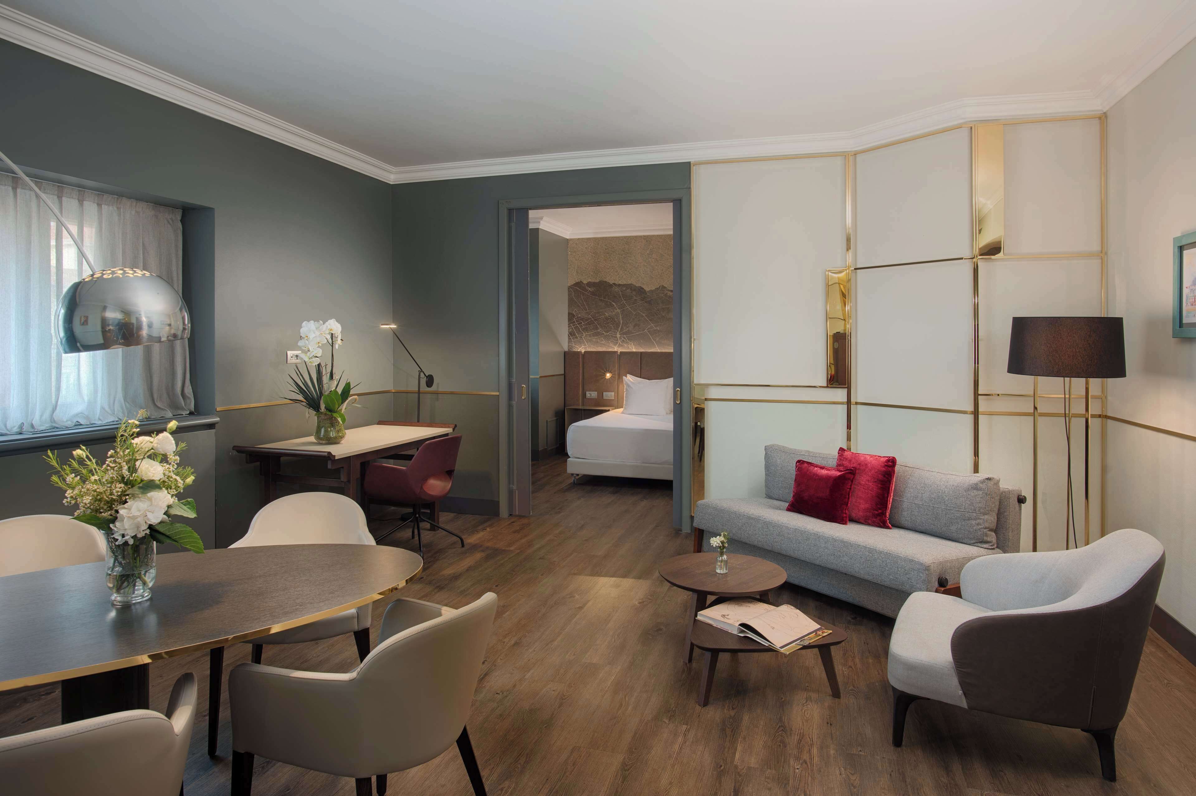 A modern hotel room with a dining area, a sofa, a bed, and a desk.