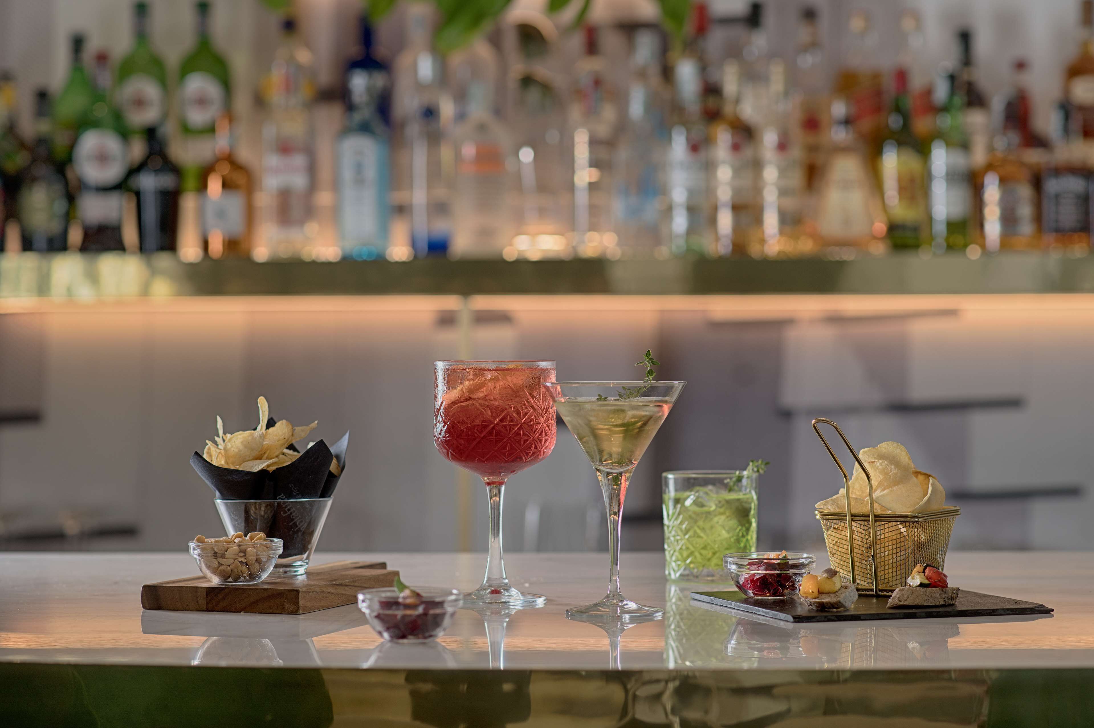 A bar area with a variety of drinks and snacks, including a martini, a cocktail with a red drink, and a green drink. There are also bowls of nuts and a basket of bread.