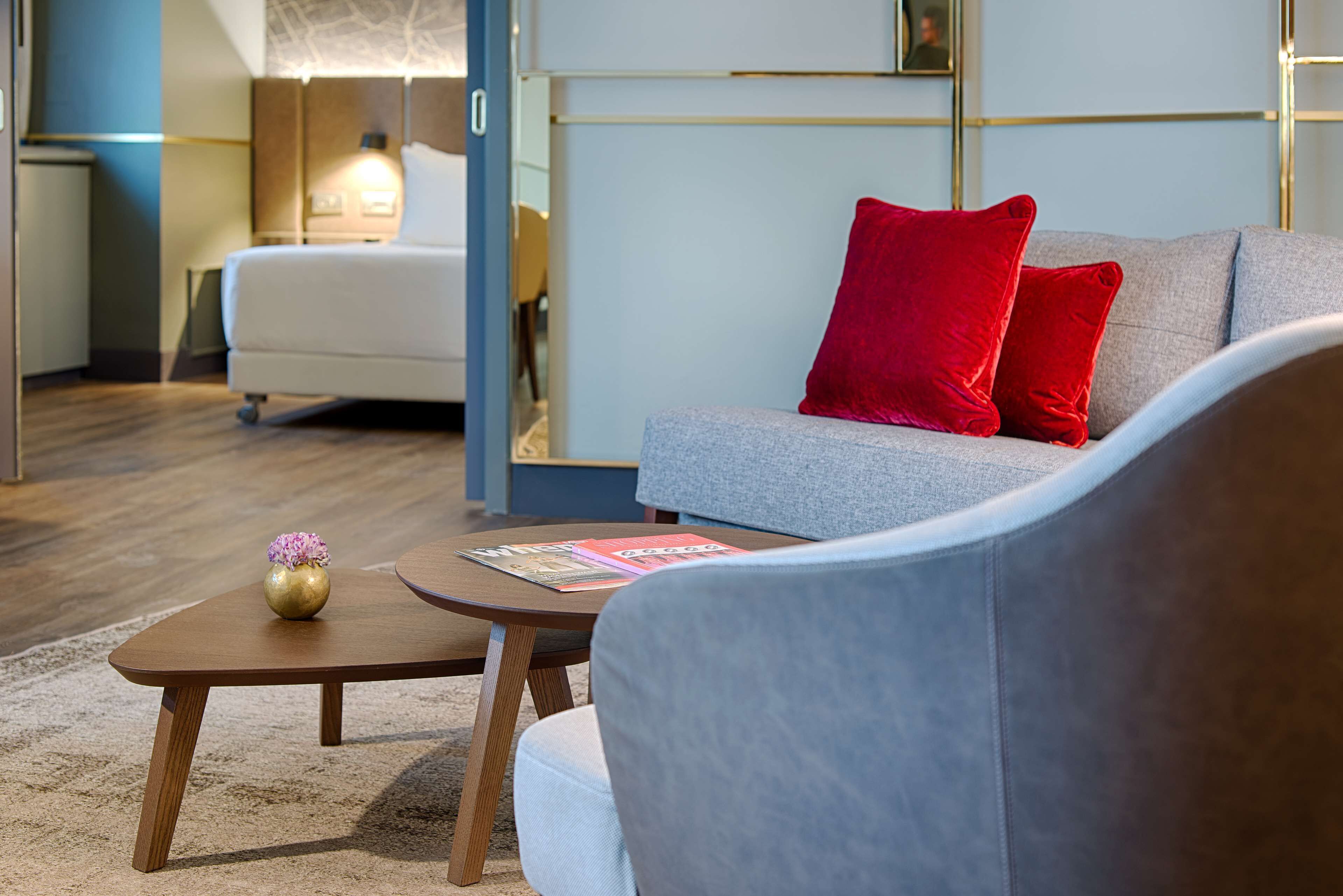 Ein modernes Wohnzimmer mit einem grauen Sofa, roten Kissen, einem Holztisch mit Vase und Zeitschriften sowie einem weißen Sessel.