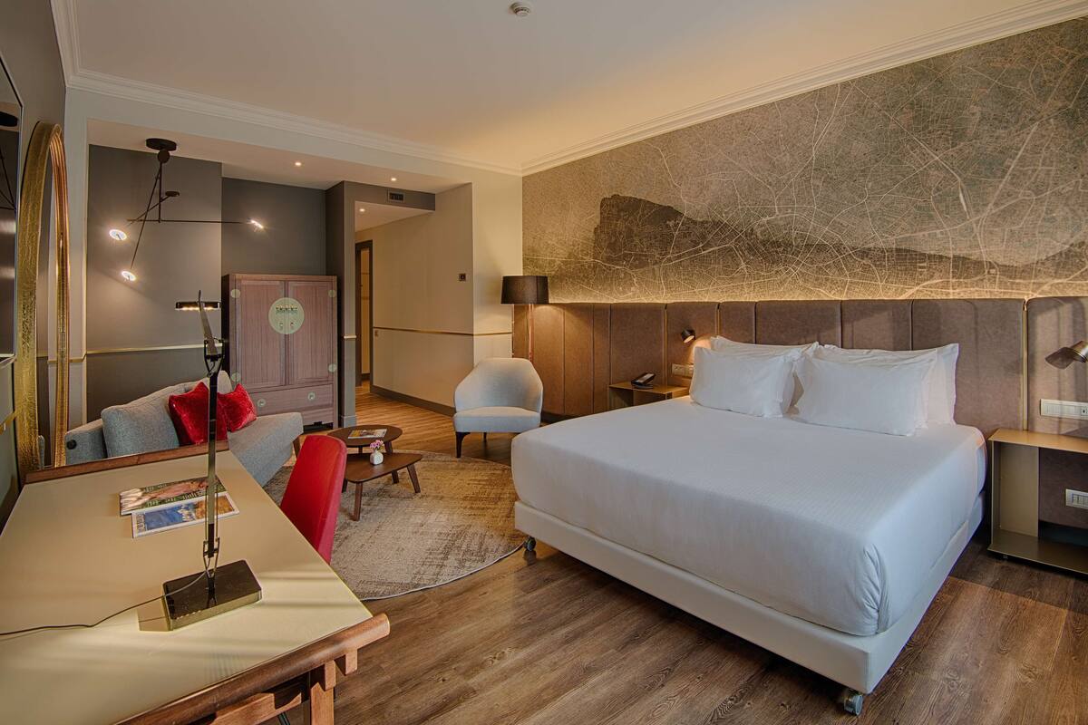 A modern hotel room with a large bed, a desk with a lamp, a chair, a map on the wall, and a mirror.