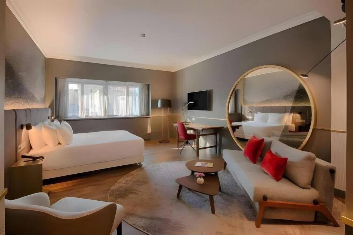 A modern hotel room with a bed, sofa, desk, and a large mirror.
