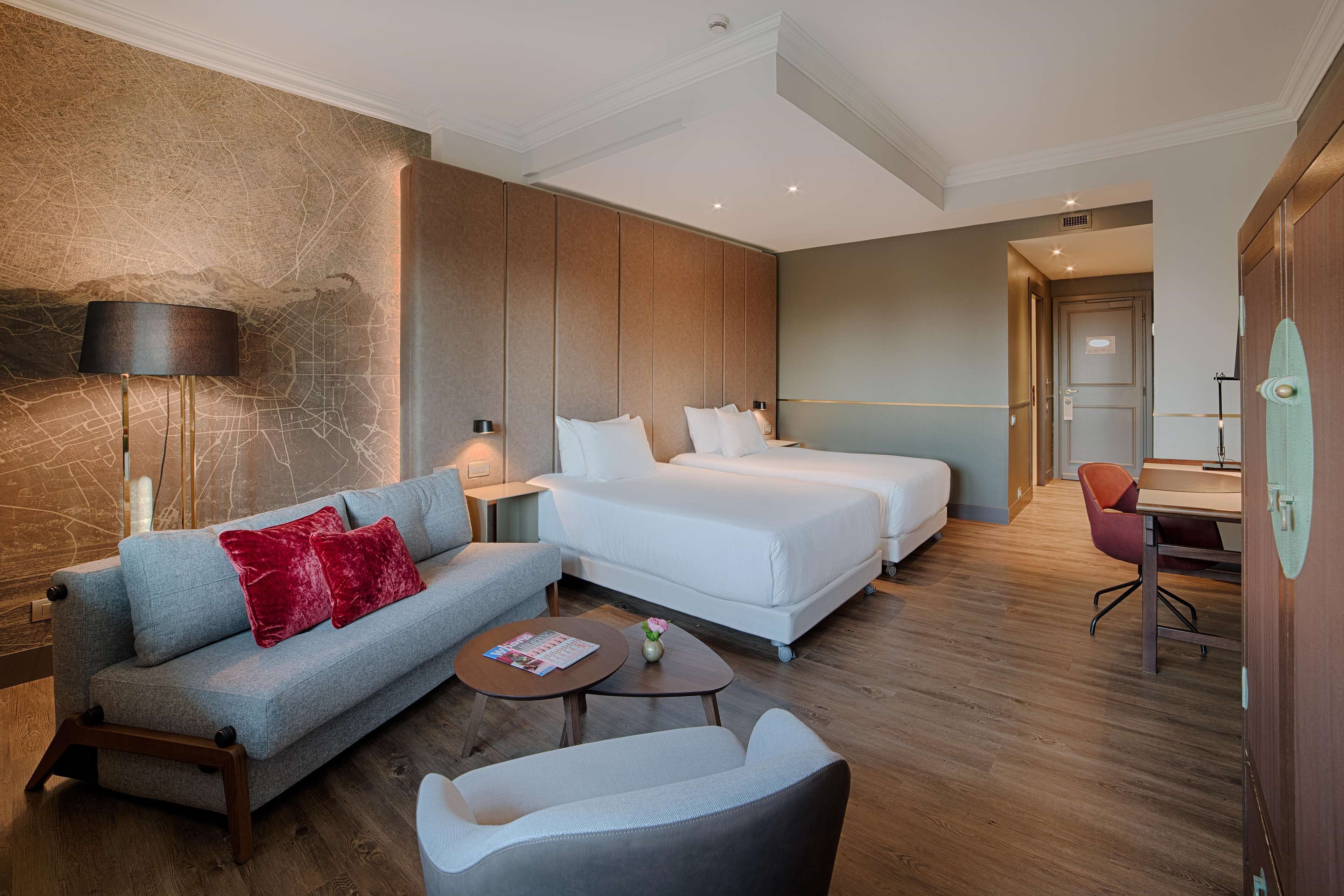 A modern hotel room with a sofa, two single beds, a desk, and a chair.