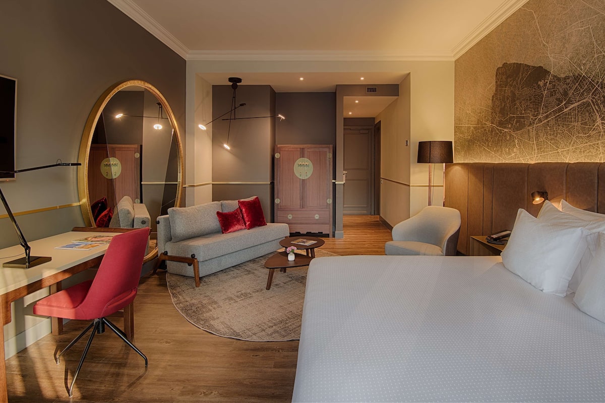 A modern hotel room with a large bed, a desk with a chair, a sofa, and a large mirror.