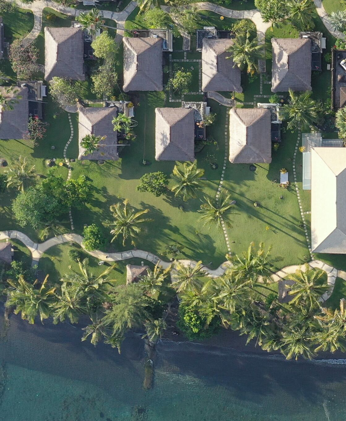 Luchtbeeld van een resort met rieten huisjes, geordend in een raster, omgeven door palmbomen en een helderblauw water.