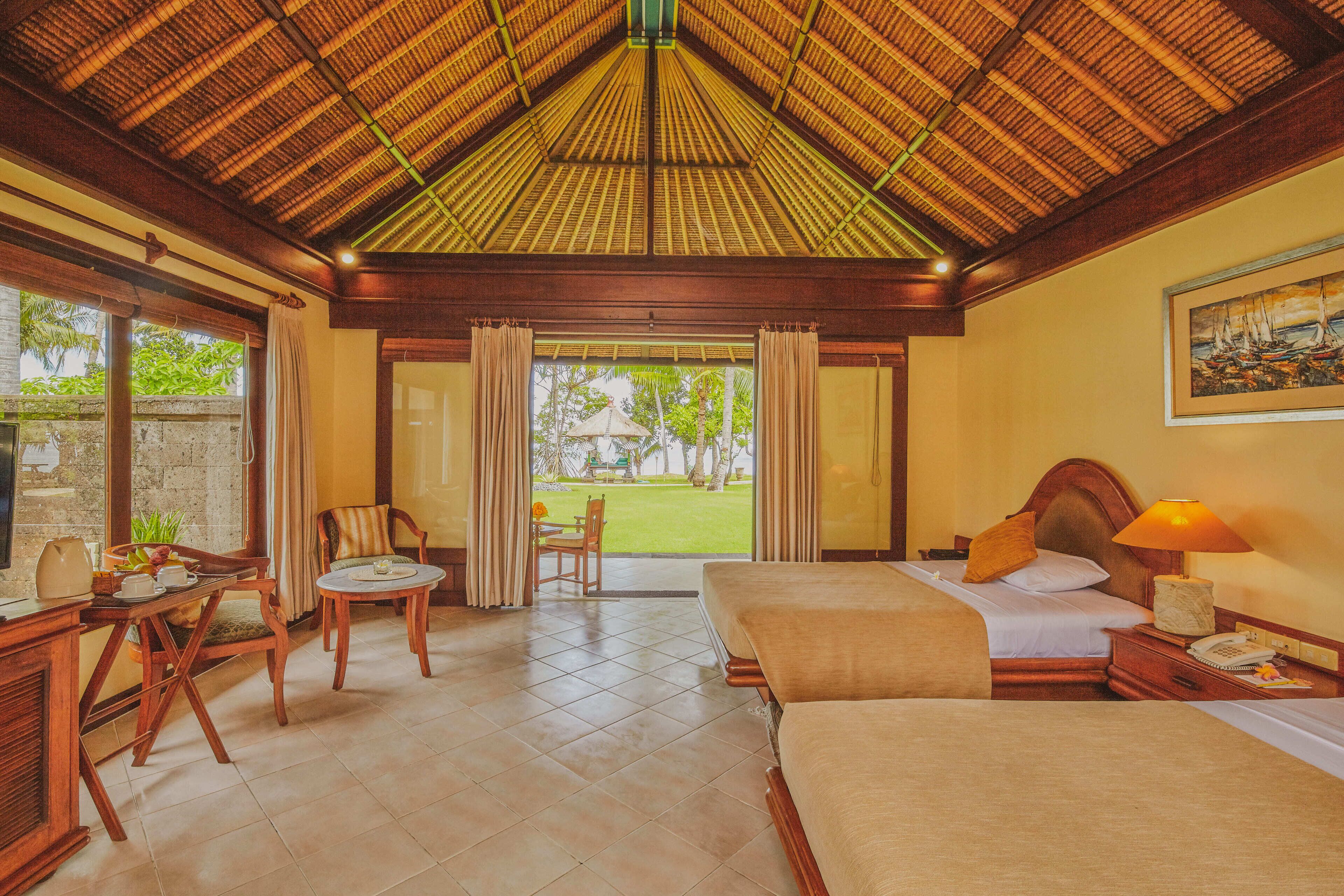 Habitación de hotel con techo de paja, una cama, un escritorio con silla y vista al jardín.