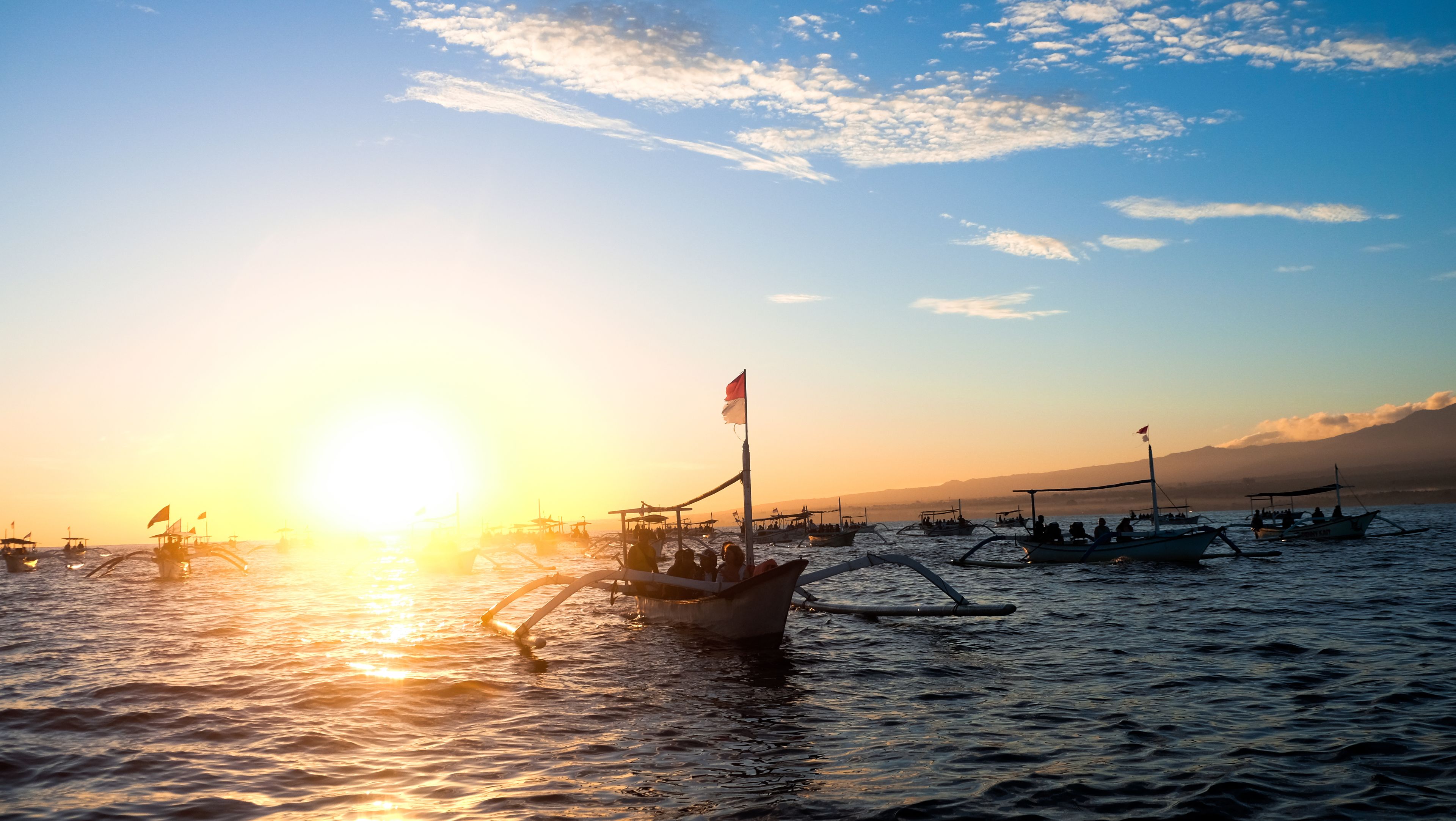 Un coucher de soleil sur une étendue d’eau, avec plusieurs bateaux et un bateau amarré portant un drapeau.