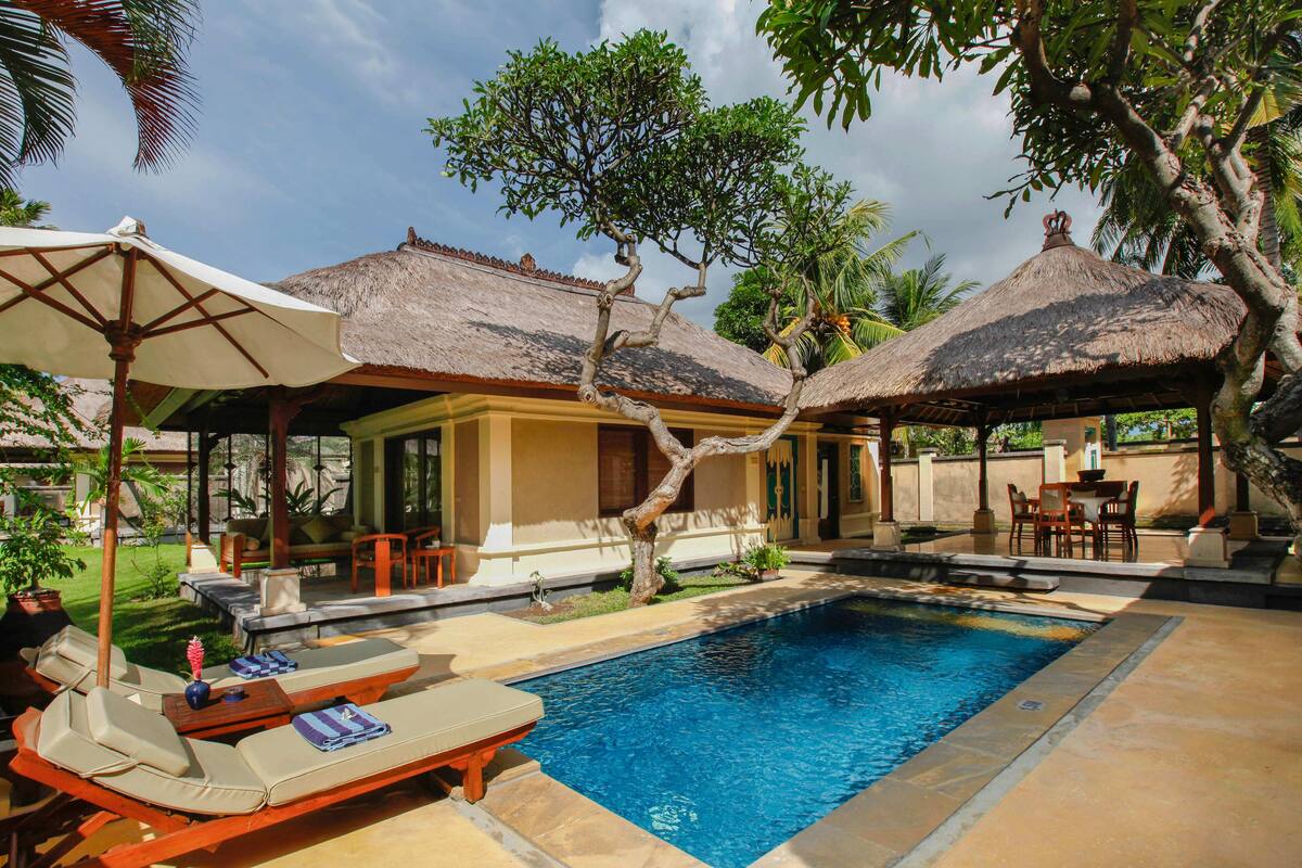 A pool area with lounge chairs, umbrellas, and a thatched-roof structure.