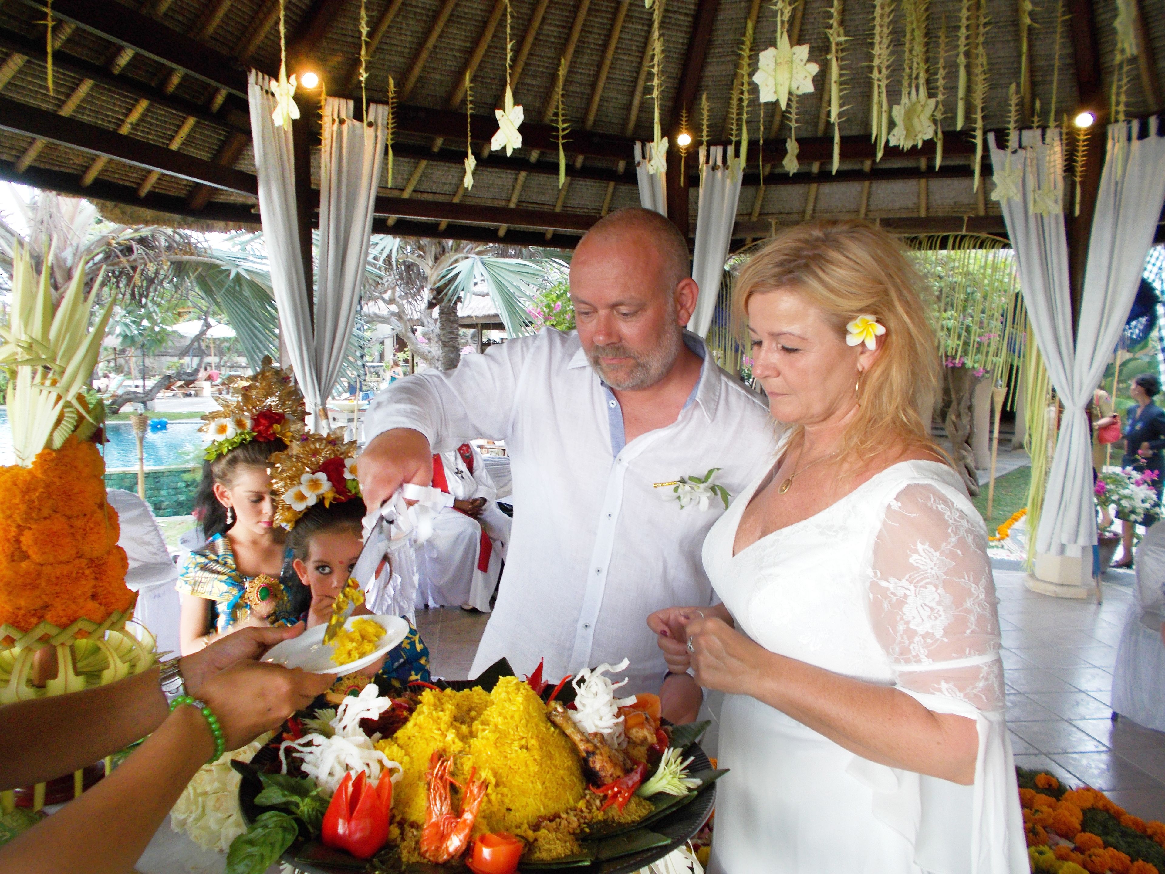 A wedding ceremony with a tropical setting, featuring a covered area with a thatched roof and hanging floral decorations.