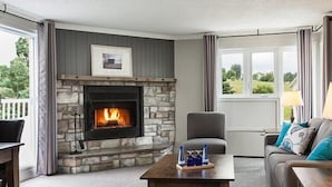 A living room with a stone fireplace, a grey couch, a wooden coffee table, and a view of the outdoors. - Auberge West Brome (Lac-Brome)