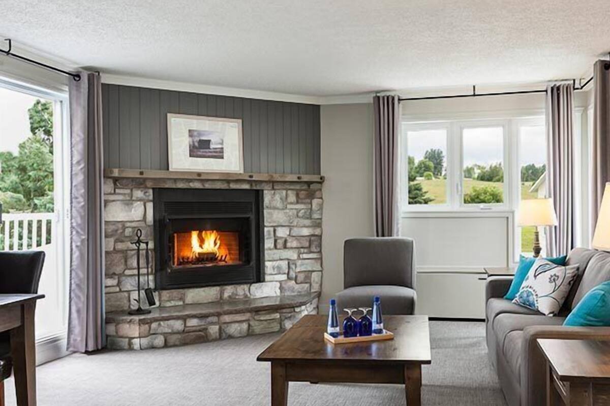 A living room with a stone fireplace, a grey couch, a wooden coffee table, and a view of the outdoors.