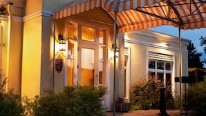A hotel entrance with a covered patio, a well-lit facade, and a landscaped garden area. - Auberge West Brome (Lac-Brome)