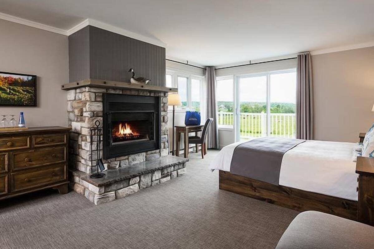 A bedroom with a stone fireplace, a bed, a desk, and a balcony with a view.