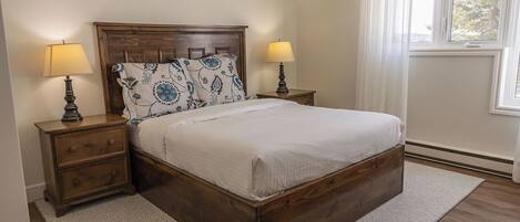 Un dormitorio con una cama de madera, dos mesitas de noche con lámparas, un cojín estampado y una ventana con cortinas.