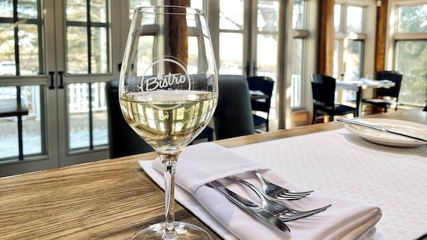 A glass of white wine on a wooden table with a napkin and fork, in a room with large windows and wooden beams. - Auberge West Brome (Lac-Brome)